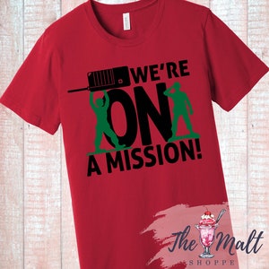 May include: Red t-shirt with a black and green graphic design. The graphic features two toy soldiers holding a walkie-talkie and the text "We're On A Mission!"
