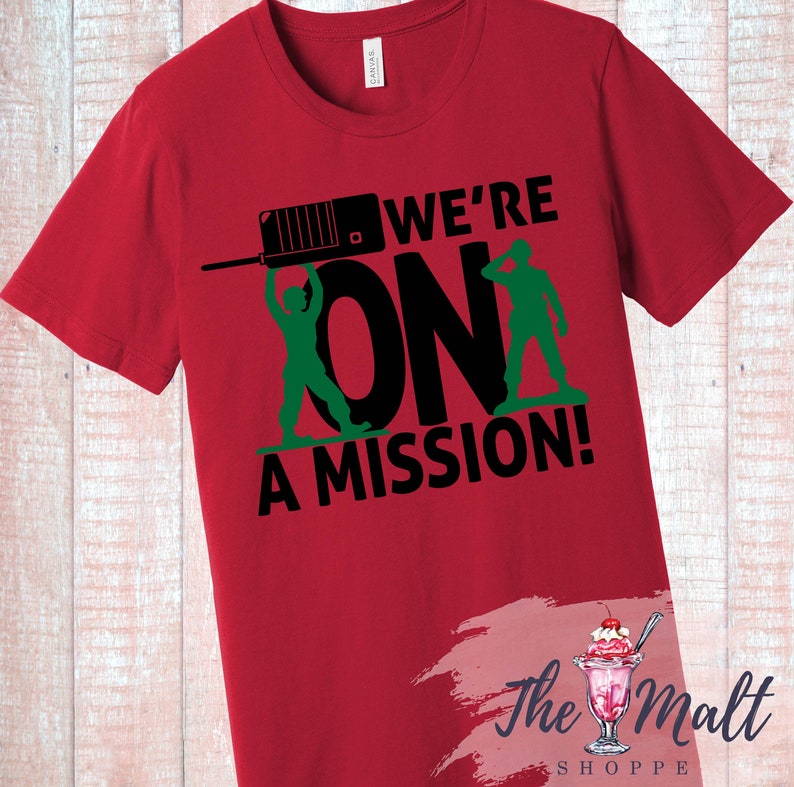 May include: Red t-shirt with a black and green graphic design. The graphic features two toy soldiers holding a walkie-talkie and the text "We're On A Mission!"