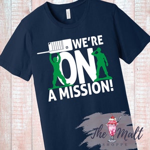 May include: Navy blue t-shirt with a white graphic of two toy soldiers holding up the word "ON" with the text "WE'RE ON A MISSION!" above the graphic.