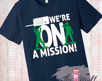 Op de afbeelding: Donkerblauw T-shirt met een witte afbeelding van twee speelgoed soldaten die het woord "ON" omhoog houden met de tekst "WE'RE ON A MISSION!" boven de afbeelding.