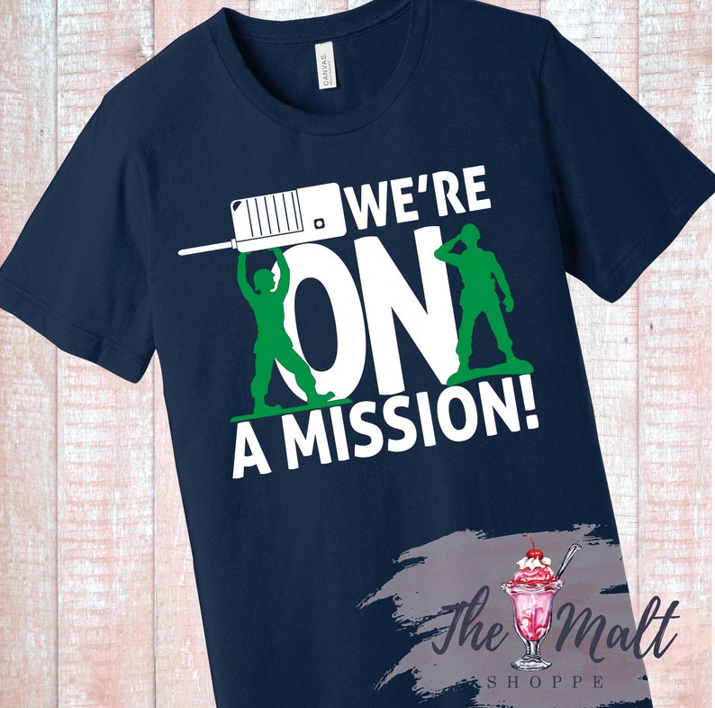 May include: Navy blue t-shirt with a white graphic of two toy soldiers holding up the word "ON" with the text "WE'RE ON A MISSION!" above the graphic.