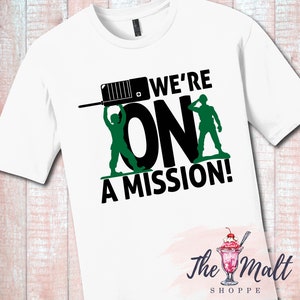 May include: White t-shirt with a black and green graphic design. The graphic features two toy soldiers holding up the word "ON" with a walkie-talkie above them. The text "WE'RE" is above the soldiers and the text "A MISSION!" is below them. The text "The Malt Shoppe" is in the bottom right corner of the image.