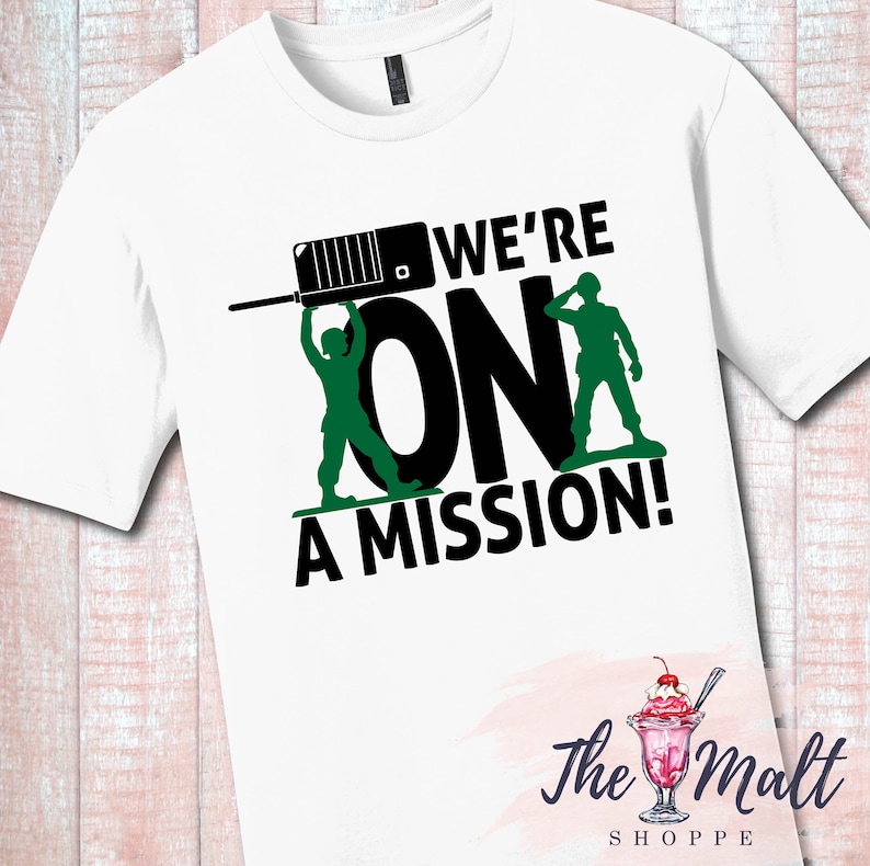 May include: White t-shirt with a black and green graphic design. The graphic features two toy soldiers holding up the word "ON" with a walkie-talkie above them. The text "WE'RE" is above the soldiers and the text "A MISSION!" is below them. The text "The Malt Shoppe" is in the bottom right corner of the image.