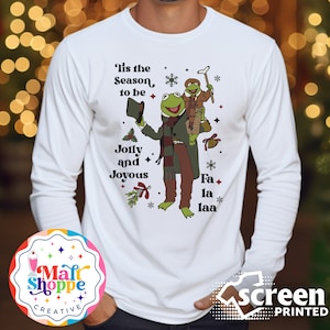 May include: White long-sleeve t-shirt with a green and brown illustration of Kermit the Frog and Miss Piggy dressed in Christmas attire. The text "Tis the Season to be Jolly and Joyous" and "Fa la laa" are printed on the shirt.
