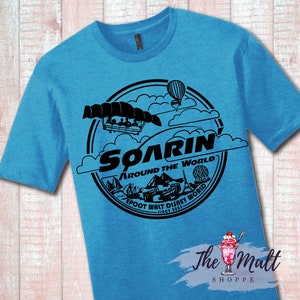 May include: A blue t-shirt with a black graphic of the Soarin' Around the World ride at Epcot. The graphic includes a hot air balloon, clouds, and the ride vehicle. The text "Soarin' Around the World Epcot Walt Disney World Since 1982" is included in the graphic.