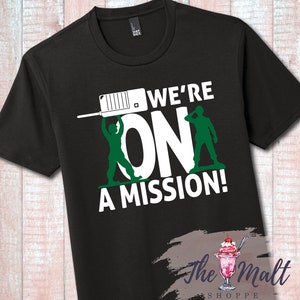 May include: Black t-shirt with a white graphic of two green toy soldiers holding a walkie-talkie and the text "We're On A Mission!"