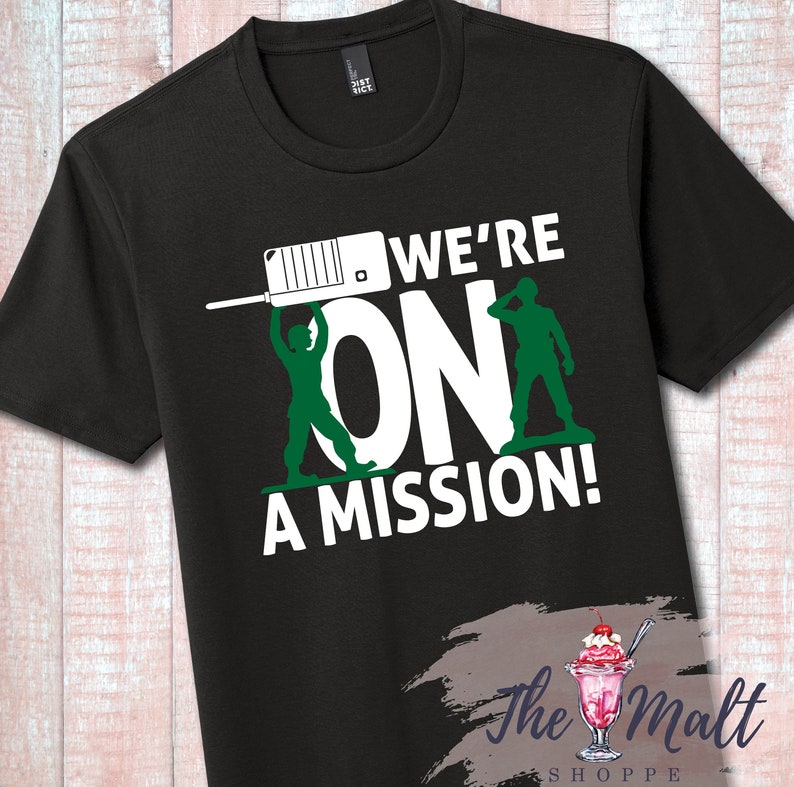 May include: Black t-shirt with a white graphic of two green toy soldiers holding a walkie-talkie and the text "We're On A Mission!"