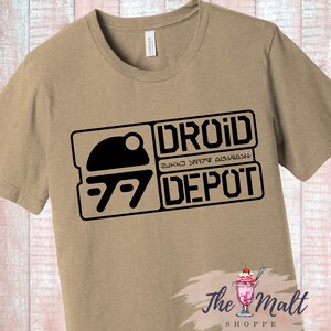 May include: A tan t-shirt with a black graphic of a droid depot logo. The logo features a stylized image of a droid head and the text "Droid Depot" in black.