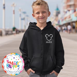 May include: A black zip-up hoodie with a white Mickey Mouse head outline and the name "JACK" printed below. The hoodie has a hood and two front pockets. The background shows a boardwalk and blurred buildings.