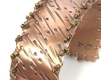Peut inclure: Un bracelet manchette en cuivre avec une surface texturée et un motif découpé décoratif. Les bords sont ornés de petites perles dorées et irisées. Le bracelet est de style large, avec les mots "Jewels of a Queen" en bas à droite.