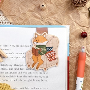 Könnte beinhalten: Ein verspielter Lesezeichen mit einem Fuchs, der in einem Sessel liest, in einem offenen Buch. Das Bild zeigt eine Rolle Washi-Tape, eine Holzklammer und einen Stift. Der Hintergrund ist braunes Papier, was eine warme Atmosphäre schafft.
