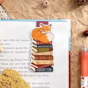 Könnte beinhalten: Ein skurriles Lesezeichen mit einem Fuchs mit Brille, der auf einem Bücherstapel ruht. Das Lesezeichen befindet sich in einem offenen Buch, mit einem Stift und einer Washi-Tape-Rolle. Auf der Washi-Tape-Rolle steht "Anna lunat" und "GINGHAM washi tape".
