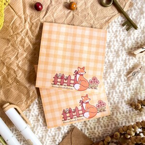May include: Two orange and white fox illustrations on a set of orange and white gingham notepad paper. The foxes are standing by a red picket fence with pink flowers. The text "@annaturaldraws" is at the bottom of each notepad.