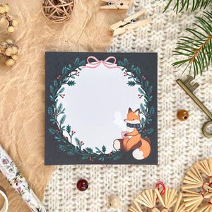 Puede incluir: Una tarjeta de felicitación cuadrada con una ilustración de un zorro, rodeada por una corona. El zorro lleva una bufanda y sostiene una taza. Otros elementos incluyen un bolígrafo, una llave y elementos decorativos sobre una superficie texturizada.