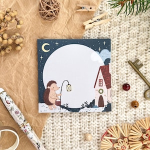 May include: A square notepad featuring a charming illustration of a hedgehog with a lantern, set against a moonlit sky and a house. The image is complemented by a pen, key, and ornaments, creating a warm, festive atmosphere.
