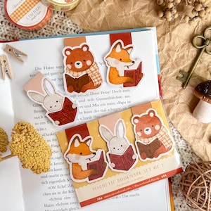 Könnte beinhalten: Eine Sammlung von herbstlichen Schreibwaren und Accessoires. Enthält dekorative Washi-Tape-Rollen, Holzklammern, einen Vintage-Schlüssel und ein Set magnetischer Lesezeichen mit Illustrationen eines Bären, Fuchses und Kaninchens, die Bücher lesen. Die Artikel sind auf einer braunen Papieroberfläche angeordnet.