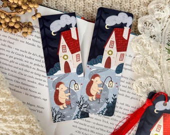 May include: Bookmarks with a whimsical illustration of a hedgehog carrying a lantern in front of a house with a red roof. The bookmarks have a night sky background with a crescent moon and clouds. The bookmarks are on top of an open book.