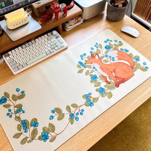 以下が含まれることがあります： オレンジ色の毛皮とふわふわの白い尻尾を持つキツネの水彩イラストが描かれた長方形の白いデスクマット。青いベリーと緑の葉に囲まれています。キーボードとマウスを収納できるように設計されています。