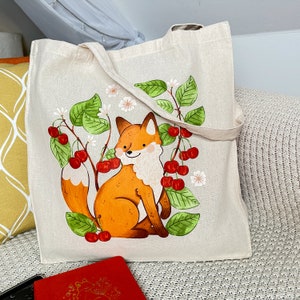 Könnte beinhalten: Beige Canvas-Tragetasche mit einer skurrilen Illustration eines Fuchses, umgeben von Kirschen und grünen Blättern. Der Fuchs ist orange und weiß mit einem lächelnden Gesichtsausdruck. Die Tasche hat beige Griffe.