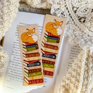 Könnte beinhalten: Ein Lesezeichen mit einem Fuchs, der sich auf einen Stapel bunter Bücher zusammengerollt hat. Der Fuchs ist orange mit einer weißen Brust und einer schwarzen Nase. Die Bücher haben unterschiedlich farbige Rücken und sind zu einem Stapel gestapelt.