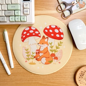 Könnte beinhalten: Rundes beiges Mauspad mit einem Cartoon-Fuchs, der zwischen zwei rot-weiß gepunkteten Pilzen sitzt. Der Fuchs hält eine Tasse. Das Mauspad hat einen cremefarbenen Rand und ist von grünem Laub und kleinen Pilzen umgeben.