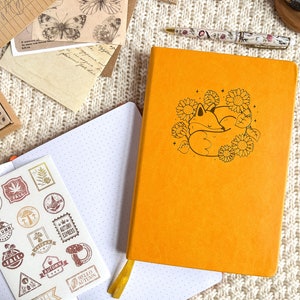 Könnte beinhalten: Ein gelbes Notizbuch mit einer schwarzen Linienzeichnung eines schlafenden Fuchses in einem Kreis aus Sonnenblumen. Das Notizbuch ist auf einer leeren Seite mit weißen Punkten geöffnet.