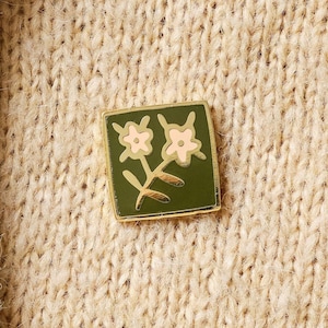 May include: A gold enamel pin with a green background featuring two pink flowers with white centres and a gold stem.