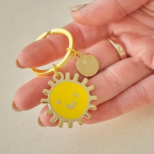Könnte beinhalten: Ein goldfarbener Schlüsselanhänger mit einem gelben Sonnenanhänger, der ein lächelndes Gesicht zeigt. Der Sonnenanhänger hat einen goldenen Umriss und ist an einem goldenen Ring befestigt.