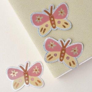 May include: Three pink and beige butterfly stickers with glitter accents. The butterflies have a whimsical, hand-drawn style.