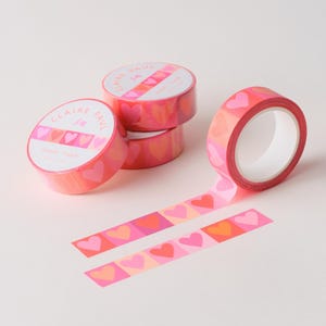May include: Pink and red heart-patterned washi tape rolls with the text "Claire Paul". One roll is unrolled, revealing a strip of tape with heart designs. The tape is ideal for crafts and adding a decorative touch.