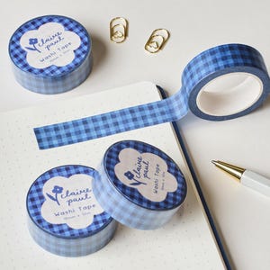 May include: Blue gingham washi tape rolls with a floral design and the text "claire paul washi tape". The image also includes gold paper clips, a pen, and a notebook. The tape is 15mm x 10m.