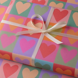 May include: A present wrapped in paper patterned with colourful hearts in pink, orange, red, green, and purple. A cream ribbon is tied around the gift in a bow.