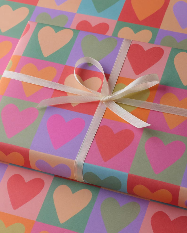 May include: A present wrapped in paper patterned with colourful hearts in pink, orange, red, green, and purple. A cream ribbon is tied around the gift in a bow.