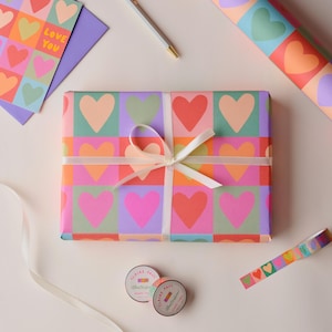 May include: A gift wrapped in paper with a vibrant heart design, secured with a white ribbon. Coordinated stationery, including a card with the words "Love You", a pen, and decorative tape, are also present. The design features hearts in various colours.