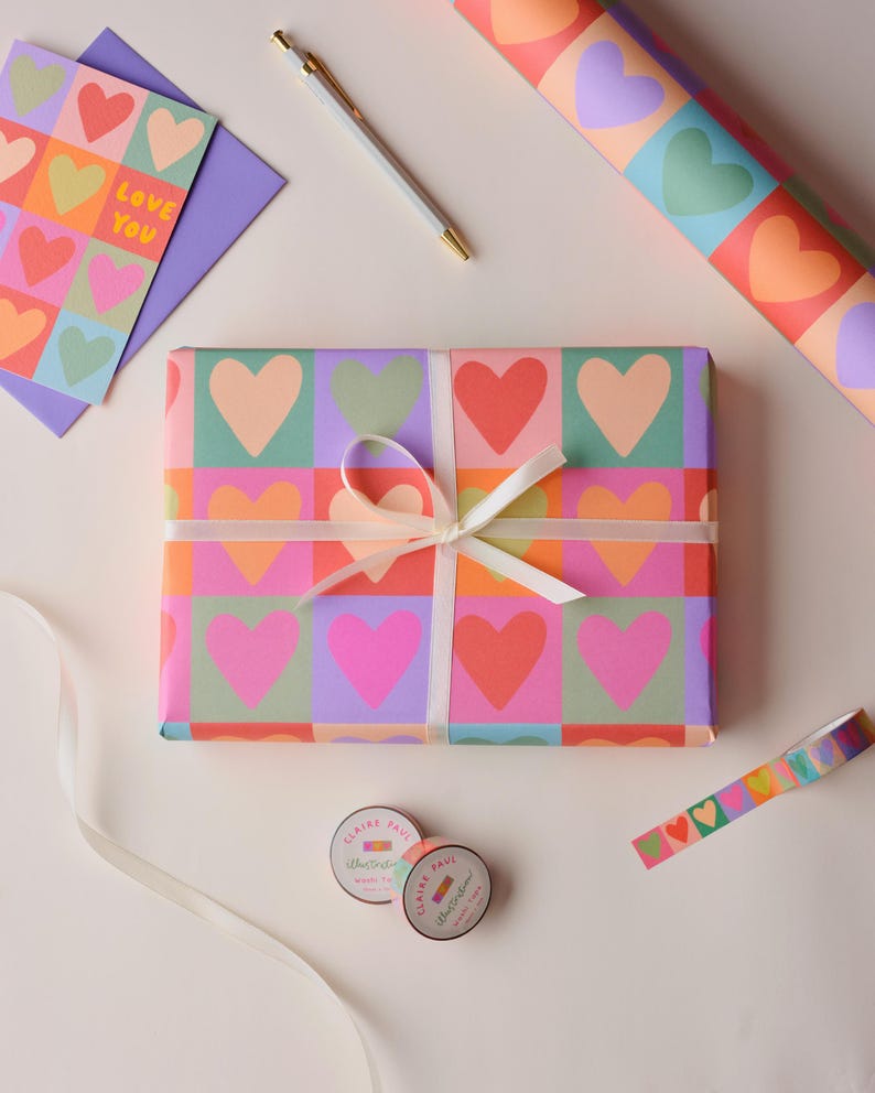 May include: A gift wrapped in paper with a vibrant heart design, secured with a white ribbon. Coordinated stationery, including a card with the words "Love You", a pen, and decorative tape, are also present. The design features hearts in various colours.