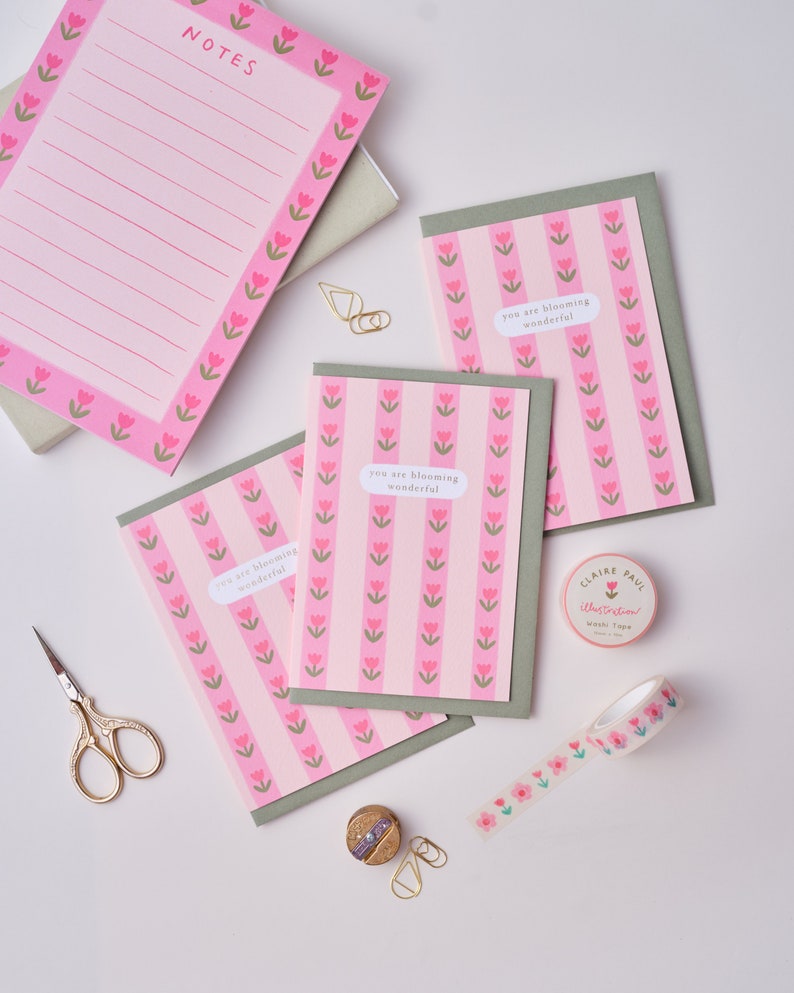 May include: Three pink and green greeting cards with the text "you are blooming wonderful" on each card. The cards are surrounded by gold paper clips, a small spool of pink and green floral washi tape, and a pair of gold scissors.