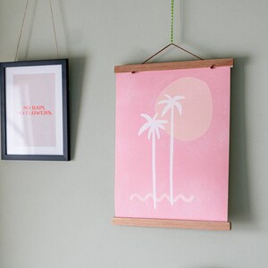 May include: Two framed prints hanging on a light green wall. The print on the left is black framed and features the text "NO RAIN, NO FLOWERS" in red. The print on the right is a pink canvas print with a white wooden frame. The print features two white palm trees and a pink sun.