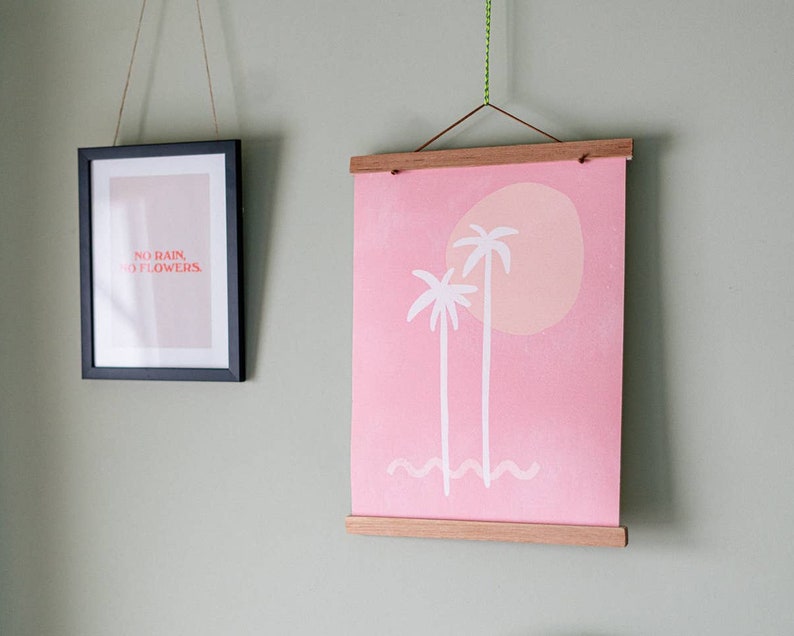 May include: Two framed prints hanging on a light green wall. The print on the left is black framed and features the text "NO RAIN, NO FLOWERS" in red. The print on the right is a pink canvas print with a white wooden frame. The print features two white palm trees and a pink sun.