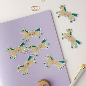 May include: Six gold foil horse stickers with green and pink details on a purple background. The stickers are arranged in a semi-circle around a gold pencil sharpener. The text "Made in Japan" is visible on the background.