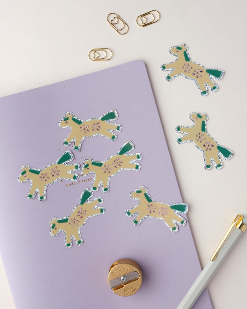 May include: Six gold foil horse stickers with green and pink details on a purple background. The stickers are arranged in a semi-circle around a gold pencil sharpener. The text "Made in Japan" is visible on the background.