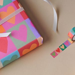 May include: A gift box wrapped in colourful paper with heart patterns, tied with a white ribbon. A roll of decorative tape with heart designs is next to the box. A pen is also visible.