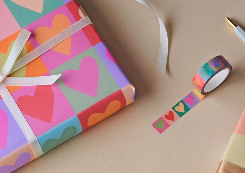 May include: A gift box wrapped in colourful paper with heart patterns, tied with a white ribbon. A roll of decorative tape with heart designs is next to the box. A pen is also visible.