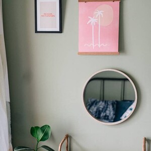 May include: A light green wall with a framed print of two palm trees on a pink background, a framed print with the text "No rain, no flowers", a round mirror with a wooden frame, and a bamboo shelf with a potted plant.