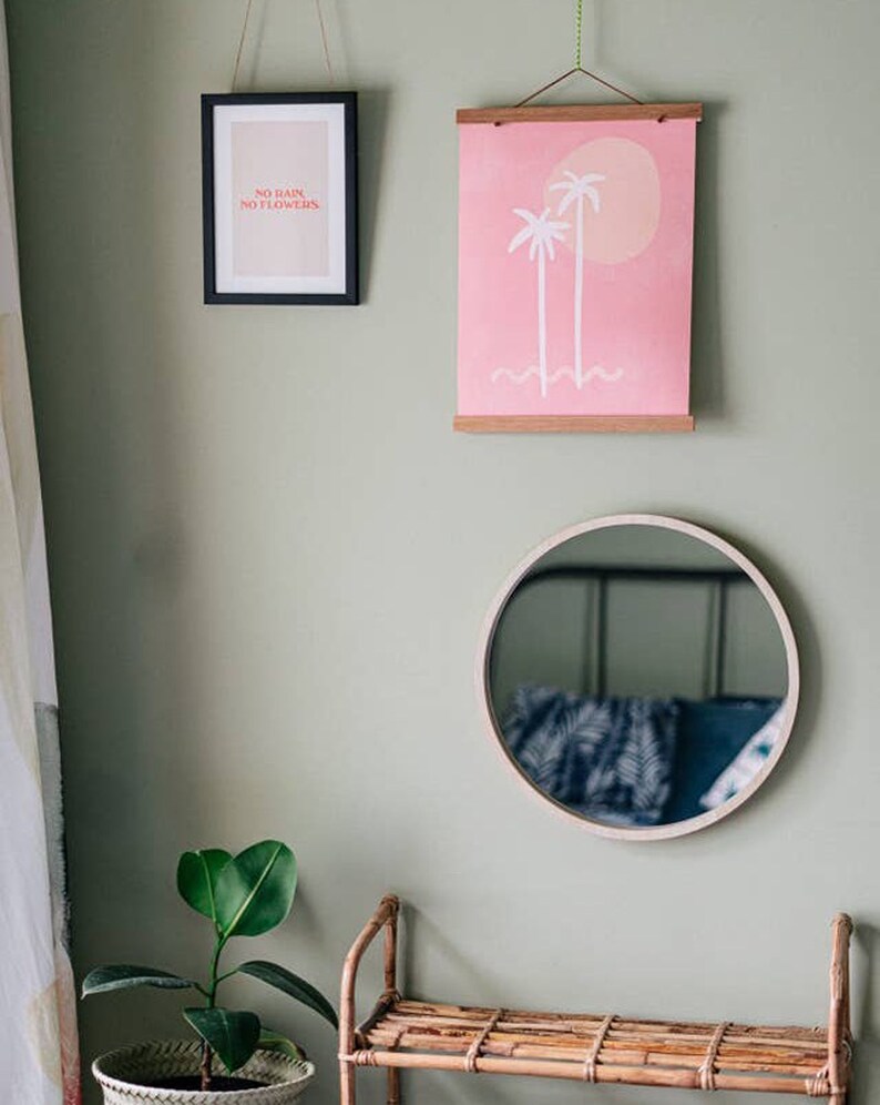 May include: A light green wall with a framed print of two palm trees on a pink background, a framed print with the text "No rain, no flowers", a round mirror with a wooden frame, and a bamboo shelf with a potted plant.