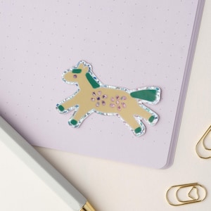 May include: A yellow horse sticker with green details and glitter accents. The sticker is on a light purple background.