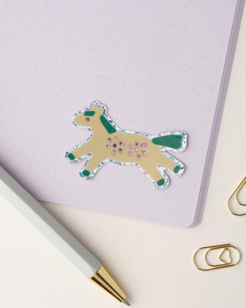 May include: A yellow horse sticker with green details and glitter accents. The sticker is on a light purple background.