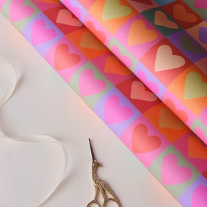 May include: Brightly coloured wrapping paper featuring a repeating heart design in pink, orange, green, and red squares. Gold scissors and a white ribbon are also visible.