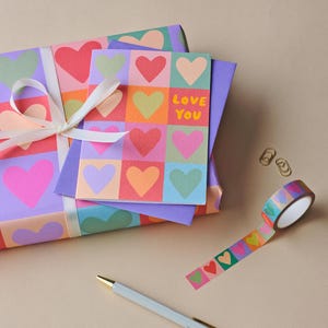 May include: A gift wrapped in paper with a heart pattern, tied with a white ribbon. A card with the words "Love You" and a heart design sits on top. A roll of heart-patterned tape, two paper clips, and a pen are also present.