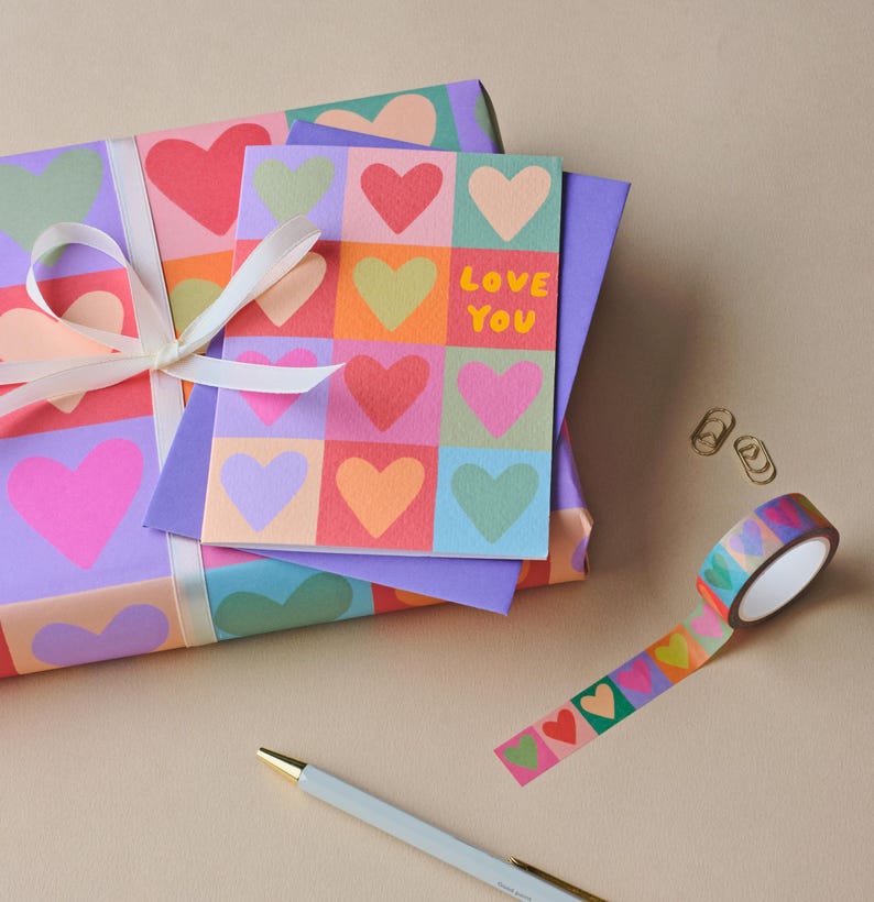 May include: A gift wrapped in paper with a heart pattern, tied with a white ribbon. A card with the words "Love You" and a heart design sits on top. A roll of heart-patterned tape, two paper clips, and a pen are also present.