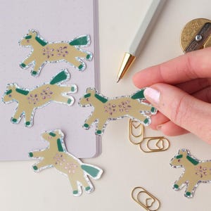 May include: Five gold glitter horse stickers with green and pink details. The stickers are on a white surface with a notebook and a pen in the background.
